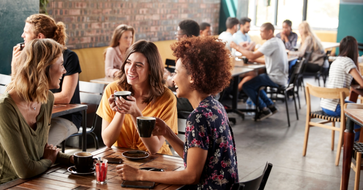 Three women having coffee at a coffeshop with people in the background having coffee